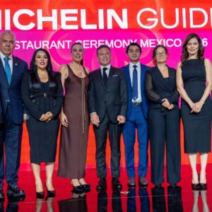 Fotografía Grupal En Un Escenario Durante La Ceremonia De La Guía Michelin México 2026. Diez Personas, Vestidas Con Trajes Formales Y Vestidos De Noche, Posan Alineadas De Pie Frente A Una Gran Pantalla Roja Brillante Que Muestra El Texto Parcial &Amp;Quot;Michelin Guide Restaurant Ceremony Mexico 2026&Amp;Quot;.