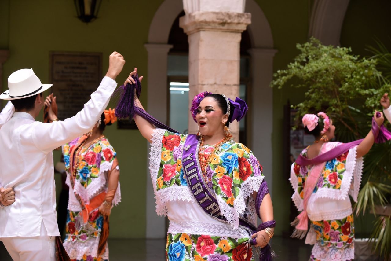 Fotografía De Un Grupo De Bailarines De Jarana Yucateca En Un Patio Colonial. En Primer Plano, Una Mujer Viste Un Terno Tradicional Blanco Con Bordados De Flores Coloridas Y Una Banda Morada Con La Palabra &Quot;Yucatán&Quot; En Letras Doradas; Sostiene Con Elegancia Sus Rebozos Mientras Baila. Detrás De Ella, Un Hombre Viste Una Guayabera Blanca Y Sombrero Panamá.