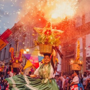 Guelaguetza 2026: presentan cartelera oficial, fechas, delegaciones confirmadas y precios de boletos la revista del sureste mx Una Fotografía Dinámica Y Colorida Capturada Al Anochecer En Una Calle Histórica De Oaxaca Durante Una Calenda. En El Centro, Una Mujer Sonriente, Vestida Con Un Traje Tradicional Que Incluye Una Falda Verde Satinada Con Encaje Blanco Y Una Blusa Bordada, Baila Mientras Equilibra En Su Cabeza Una Canasta De Mimbre Decorada Con Una Gran Estrella De Flore