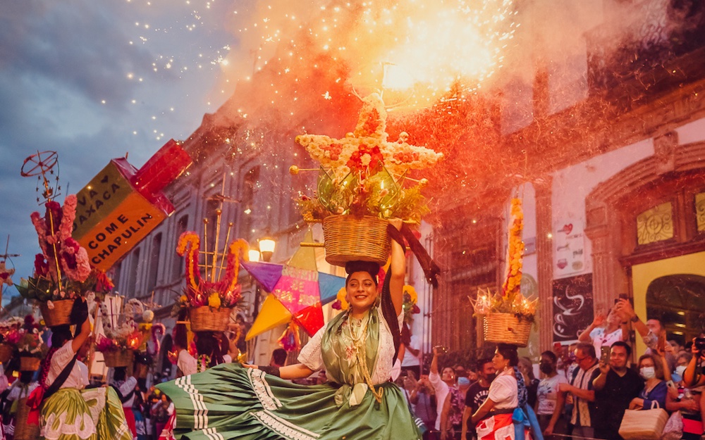 Una Fotografía Dinámica Y Colorida Capturada Al Anochecer En Una Calle Histórica De Oaxaca Durante Una Calenda. En El Centro, Una Mujer Sonriente, Vestida Con Un Traje Tradicional Que Incluye Una Falda Verde Satinada Con Encaje Blanco Y Una Blusa Bordada, Baila Mientras Equilibra En Su Cabeza Una Canasta De Mimbre Decorada Con Una Gran Estrella De Flore