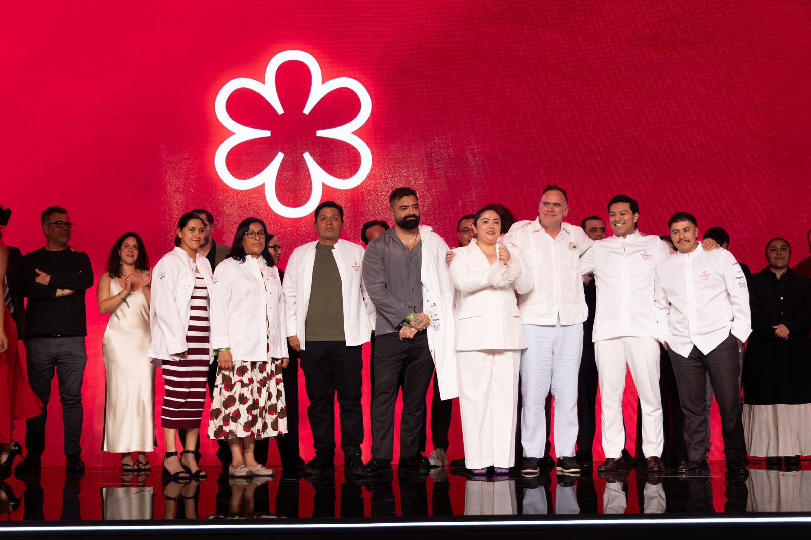 Fotografía Grupal En Un Escenario Durante Un Evento Culinario. Un Grupo De Chefs Y Profesionales De La Gastronomía, Varios De Ellos Vistiendo Filipinas Blancas De Cocina, Posan Alineados De Pie. Detrás De Ellos Destaca Una Enorme Pantalla Roja Brillante Iluminada Con El Logotipo Blanco En Forma De Flor De La Guía Michelin. El Suelo Del Escenario Es Negro Y Refleja Las Figuras De Los Asistentes.