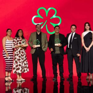 Fotografía De Plano Entero Sobre Un Escenario Con Fondo Rojo Brillante Que Muestra A Un Grupo De Seis Personas Durante La Premiación De La Guía Michelin México 2026 Yucatán. En El Centro, Dos Chefs Masculinos Sostienen Galardones De Madera Con La Silueta De La Estrella Verde Michelin En Acrílico Translúcido. A La Izquierda, Dos Mujeres Posan Sonrientes, Y A La Derecha, Un Hombre En Traje Formal Negro Y Una Mujer Con Vestido Asimétrico Oscuro Completan La Línea De Galardonados.