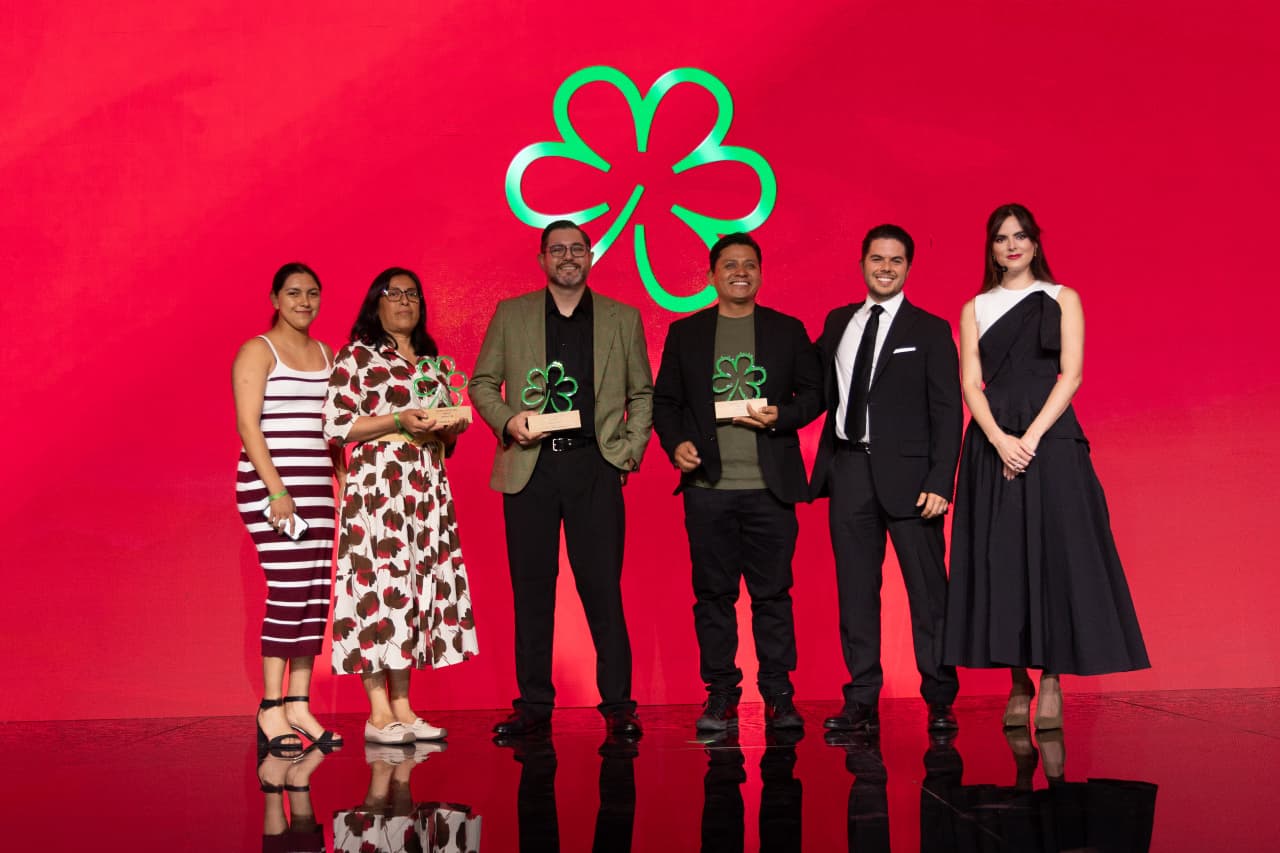 Fotografía De Plano Entero Sobre Un Escenario Con Fondo Rojo Brillante Que Muestra A Un Grupo De Seis Personas Durante La Premiación De La Guía Michelin México 2026 Yucatán. En El Centro, Dos Chefs Masculinos Sostienen Galardones De Madera Con La Silueta De La Estrella Verde Michelin En Acrílico Translúcido. A La Izquierda, Dos Mujeres Posan Sonrientes, Y A La Derecha, Un Hombre En Traje Formal Negro Y Una Mujer Con Vestido Asimétrico Oscuro Completan La Línea De Galardonados.