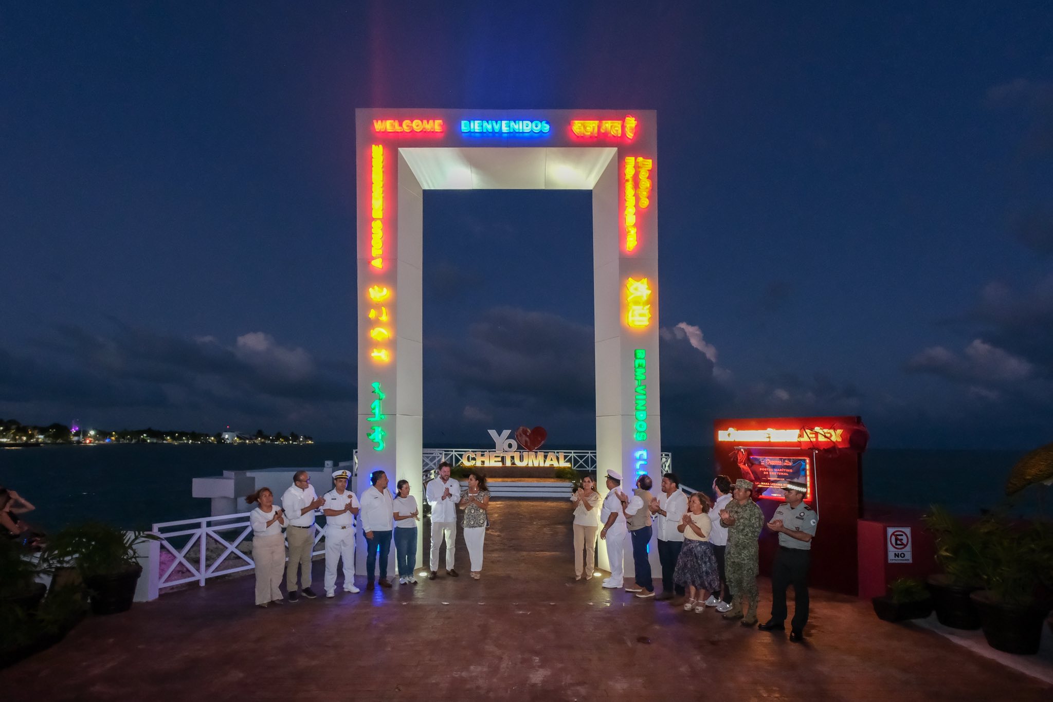 Transformación en la capital: inaugura mara lezama el portal marítimo de chetumal como nuevo ícono turístico de la bahía la revista del sureste mx Fotografía Nocturna Que Muestra A Un Grupo De Autoridades Civiles Y Militares Reunidas En El Muelle Fiscal Frente Al Mar. Al Fondo, Destaca Un Arco Monumental Iluminado Con Luces Led Multicolores Que Proyectan La Palabra &Quot;Bienvenidos&Quot; Y Otros Mensajes En Distintos Idiomas. A Través Del Arco, Se Puede Apreciar El Parador Fotográfico Con Las Letras &Quot;Yo Amo Chetumal&Quot;. El Cielo Muestra Tonalidades Oscuras De La Tarde Con Algunas Nubes Sobre La Bahía De Chetumal.