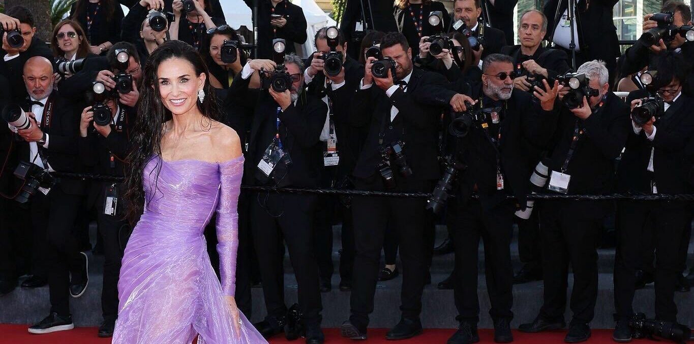 La alfombra roja de cannes 2026: entre el esplendor de la alta costura y los límites del estricto protocolo 'black tie' la revista del sureste mx Fotografía De Cuerpo Entero De La Actriz Demi Moore Posando En La Alfombra Roja Del Festival De Cannes. Viste Un Vestido De Gala Color Morado Lila Con Escote De Hombros Caídos, Mangas Largas Traslúcidas Y Una Falda Con Abertura Profunda Que Se Extiende En Una Larga Cola De Tul Vaporosa En Movimiento. Sonríe Hacia La Cámara Con El Cabello Suelto De Aspecto Húmedo. Al Fondo, Una Hilera De Fotógrafos De Prensa Con Cámaras Profesionales Captura El Momento Detrás De Una Barandilla.