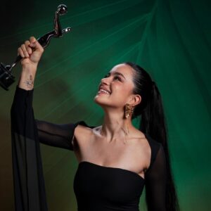 La tabasqueña paulina gaitán triunfa en los premios platino 2026: la única mexicana en conquistar la estatuilla la revista del sureste mx Fotografía De Paulina Gaitán, Vestida De Negro, Mirando Hacia Arriba Y Sosteniendo En Lo Alto El Premio Platino De Plata Sobre Un Fondo Verde Texturizado. En Su Brazo Se Aprecia Un Tatuaje Y Luce Aretes Dorados Colgantes.