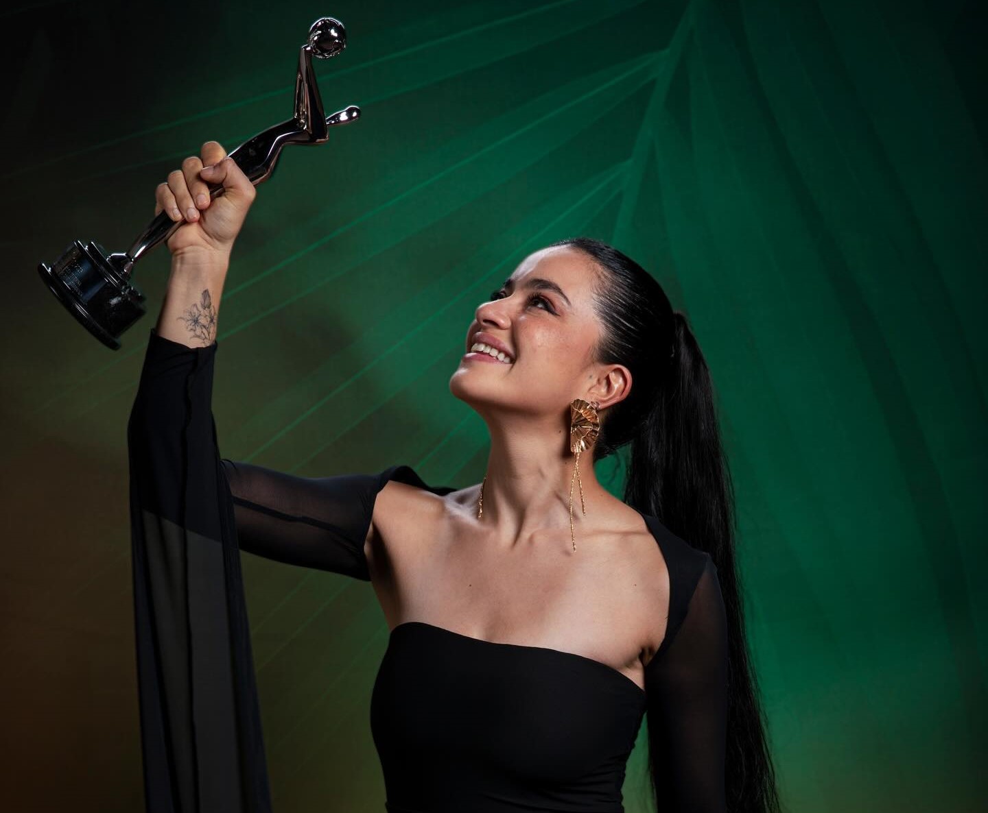 Fotografía De Paulina Gaitán, Vestida De Negro, Mirando Hacia Arriba Y Sosteniendo En Lo Alto El Premio Platino De Plata Sobre Un Fondo Verde Texturizado. En Su Brazo Se Aprecia Un Tatuaje Y Luce Aretes Dorados Colgantes.