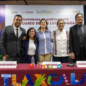 Fotografía En Plano Medio Y De Frente De Cinco Funcionarios Públicos De Pie Detrás De Una Mesa De Presídium Con Mantel Guinda Que Muestra Las Letras Coloridas De &Amp;Quot;Tlaxcala&Amp;Quot;. De Izquierda A Derecha Aparecen Un Hombre Con Traje Oscuro; La Gobernadora Lorena Cuéllar Cisneros Con Saco Azul; La Secretaria De Turismo Federal, Josefina Rodríguez Zamora.