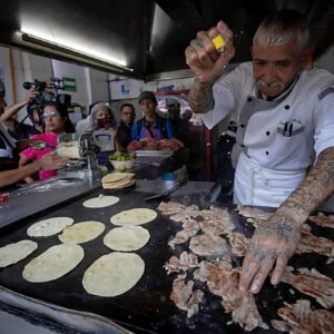¿por qué quitaron la estrella michelin a el califa de león? La revista del sureste mx Fotografía En Plano Medio De Un Parrillero De Avanzada Edad Con Tatuajes En Ambos Brazos, Trabajando En Una Taquería. Viste Un Uniforme Blanco De Chef Y Está Espolvoreando Sal Sobre Láminas De Carne Cocinándose En Un Comal Grande. A Su Lado, Una Mujer Con Cofia Blanca Prepara Tortillas. En El Fondo, Se Observa A Un Grupo De Personas Y Camarógrafos Documentando La Escena.