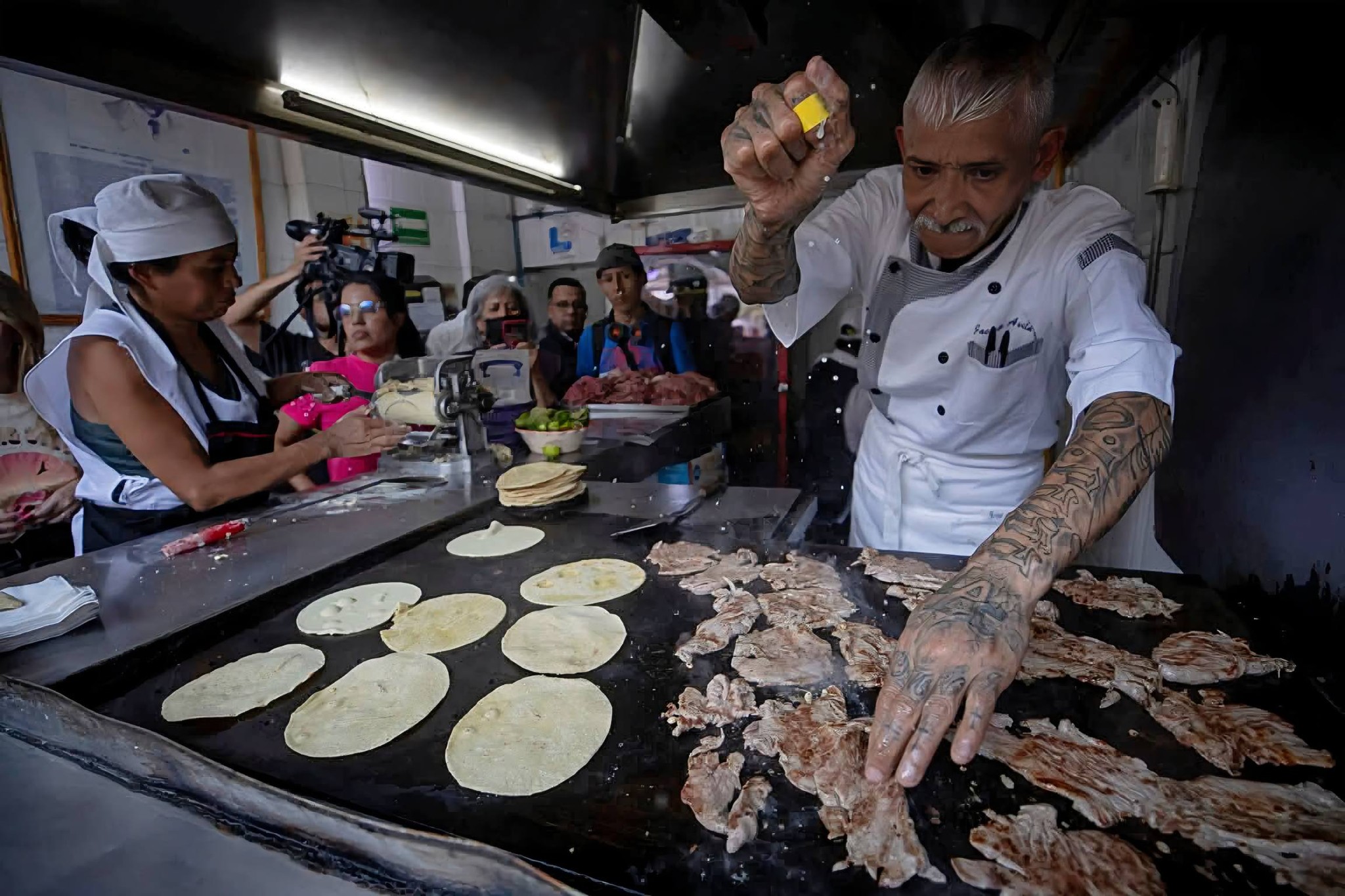Fotografía En Plano Medio De Un Parrillero De Avanzada Edad Con Tatuajes En Ambos Brazos, Trabajando En Una Taquería. Viste Un Uniforme Blanco De Chef Y Está Espolvoreando Sal Sobre Láminas De Carne Cocinándose En Un Comal Grande. A Su Lado, Una Mujer Con Cofia Blanca Prepara Tortillas. En El Fondo, Se Observa A Un Grupo De Personas Y Camarógrafos Documentando La Escena.