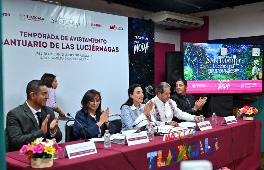 Fotografía De Ángulo Medio Tomada Desde La Derecha Que Muestra Una Conferencia De Prensa En Punto México. Una Mesa De Presídium Con Mantel Guinda Ocupa El Primer Plano. Cuatro Funcionarios Públicos Están Sentados, Todos Aplaudiendo; De Izquierda A Derecha, Un Hombre Con Traje Oscuro, La Gobernadora Lorena Cuéllar Cisneros Con Saco Azul Oscuro, La Secretaria Josefina Rodríguez Zamora Con Blusa De Rayas Y Un Hombre Con Guayabera Blanca Bordada. Detrás De Ellos, Una Gran Pancarta Blanca Con El Título &Quot;Temporada De Avistamiento Santuario De Las Luciérnagas&Quot;, Logotipos De Sectur Y Tlaxcala, Y Detalles De Fechas Y Ubicación. A La Derecha, Una Pantalla De Televisión Muestra Imágenes Del Bosque Con Luciérnagas Y El Mismo Texto Promocional. Una Asistente Está De Pie A La Izquierda.