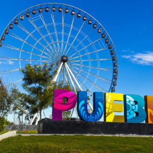 Letras Monumentales De Colores Que Forman La Palabra &Amp;Quot;Puebla&Amp;Quot; En Primer Plano, Con Diseños De Talavera Y Elementos Culturales. Al Fondo, Se Aprecia La Estrella De Puebla (Rueda De La Fortuna Gigante) Bajo Un Cielo Azul Despejado Con Nubes Ligeras.