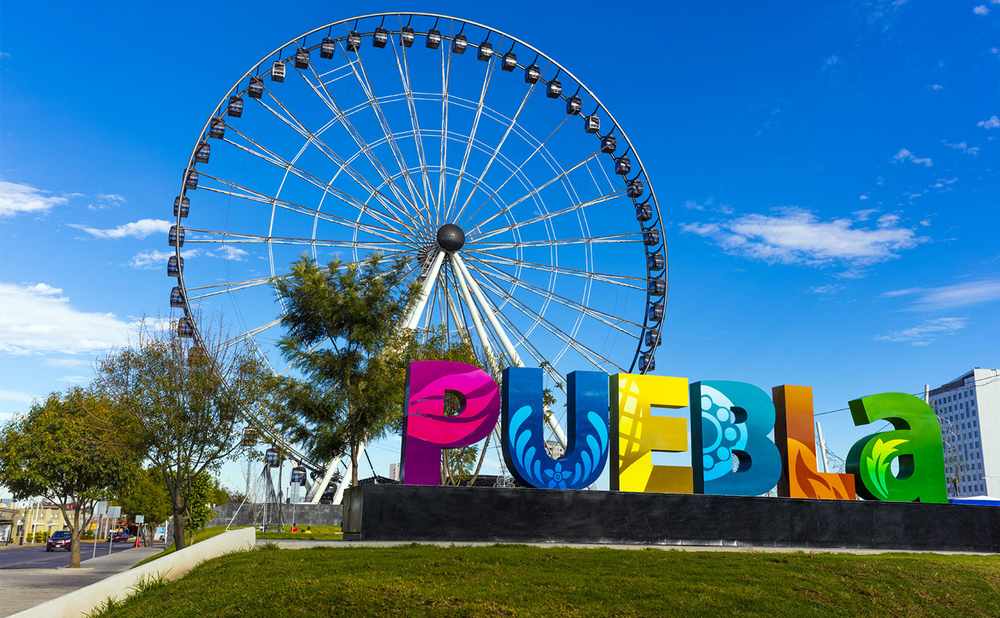 Letras Monumentales De Colores Que Forman La Palabra &Quot;Puebla&Quot; En Primer Plano, Con Diseños De Talavera Y Elementos Culturales. Al Fondo, Se Aprecia La Estrella De Puebla (Rueda De La Fortuna Gigante) Bajo Un Cielo Azul Despejado Con Nubes Ligeras.