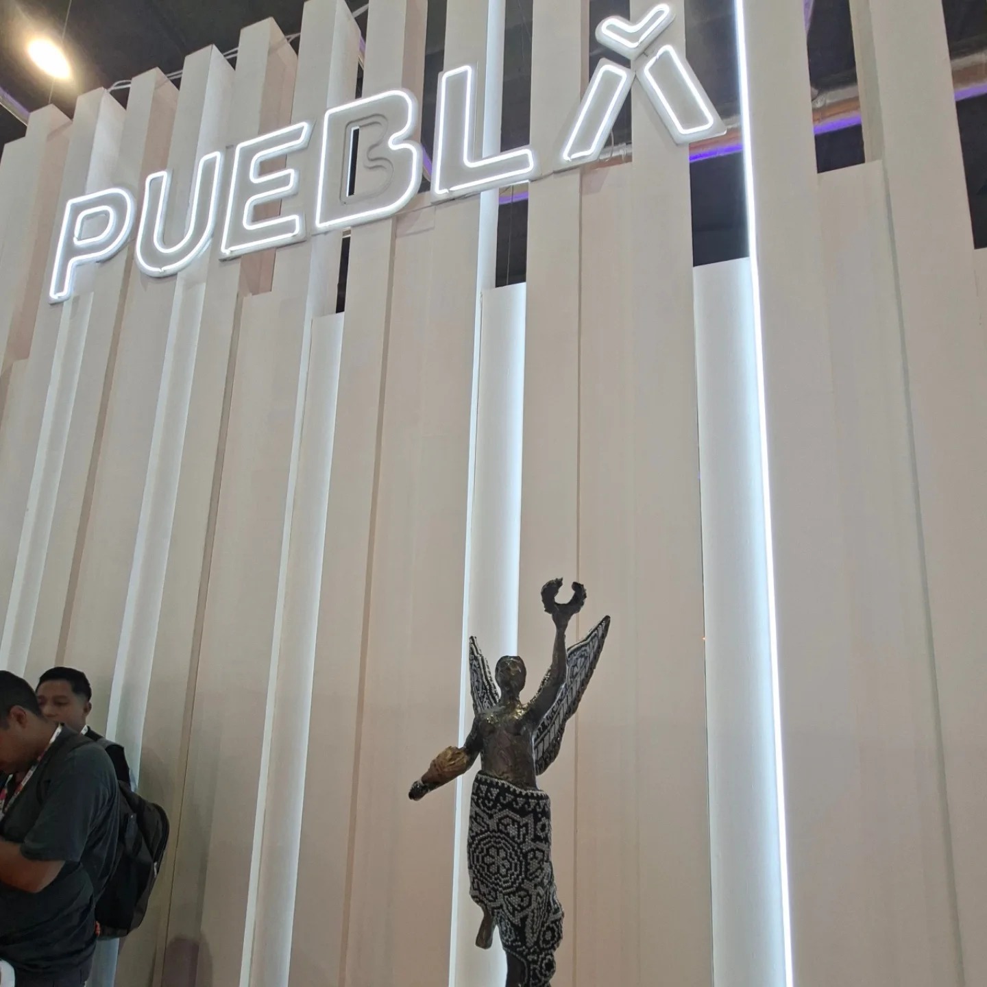 Una Estatuilla Del &Quot;Ángel Del Turismo&Quot; Con Detalles Artesanales En La Base Se Exhibe Frente A Un Muro Blanco De Paneles Verticales. En La Parte Superior Del Muro Destaca Un Letrero Luminoso Con La Palabra &Quot;Puebla&Quot; En Letras Blancas De Neón. A La Izquierda, Se Observa Parcialmente A Dos Personas En Un Entorno De Exposición.