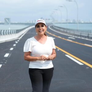 Texto Alternativo (Alt Text) La Gobernadora De Quintana Roo, Mara Lezama Espinosa, Aparece De Pie En El Centro De Uno De Los Carriles Del Nuevo Puente Vehicular Nichupté En Cancún. Viste Una Blusa Blanca, Pantalón Negro Y Una Gorra Blanca Con Logotipos Institucionales Del Estado. Detrás De Ella Se Extiende La Moderna Estructura Vial De Asfalto Impecable Con Señalización Horizontal Blanca Y Amarilla, Que Cruza Sobre El Sistema Lagunar. Al Fondo Se Aprecia El Horizonte Con La Zona Hotelera De Cancún Bajo Un Cielo Claro.