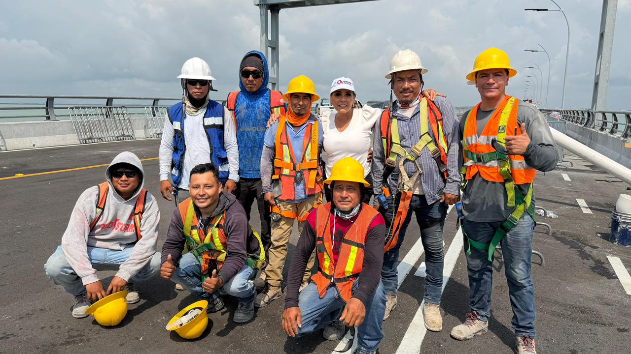 La Gobernadora Mara Lezama Posa Junto A Un Grupo De Trabajadores De La Construcción Sobre La Superficie De Rodamiento Del Puente Vehicular Nichupté. La Mandataria, Al Centro, Viste Ropa Clara Y Gorra Oficial, Mientras Que Los Trabajadores Lucen Equipo De Seguridad Que Incluye Cascos Amarillos Y Blancos, Chalecos Reflejantes Naranja Y Arneses De Protección. Al Fondo Se Observa La Estructura Metálica Del Puente, Las Barreras Laterales Y El Cielo Despejado Sobre La Laguna De Cancún.