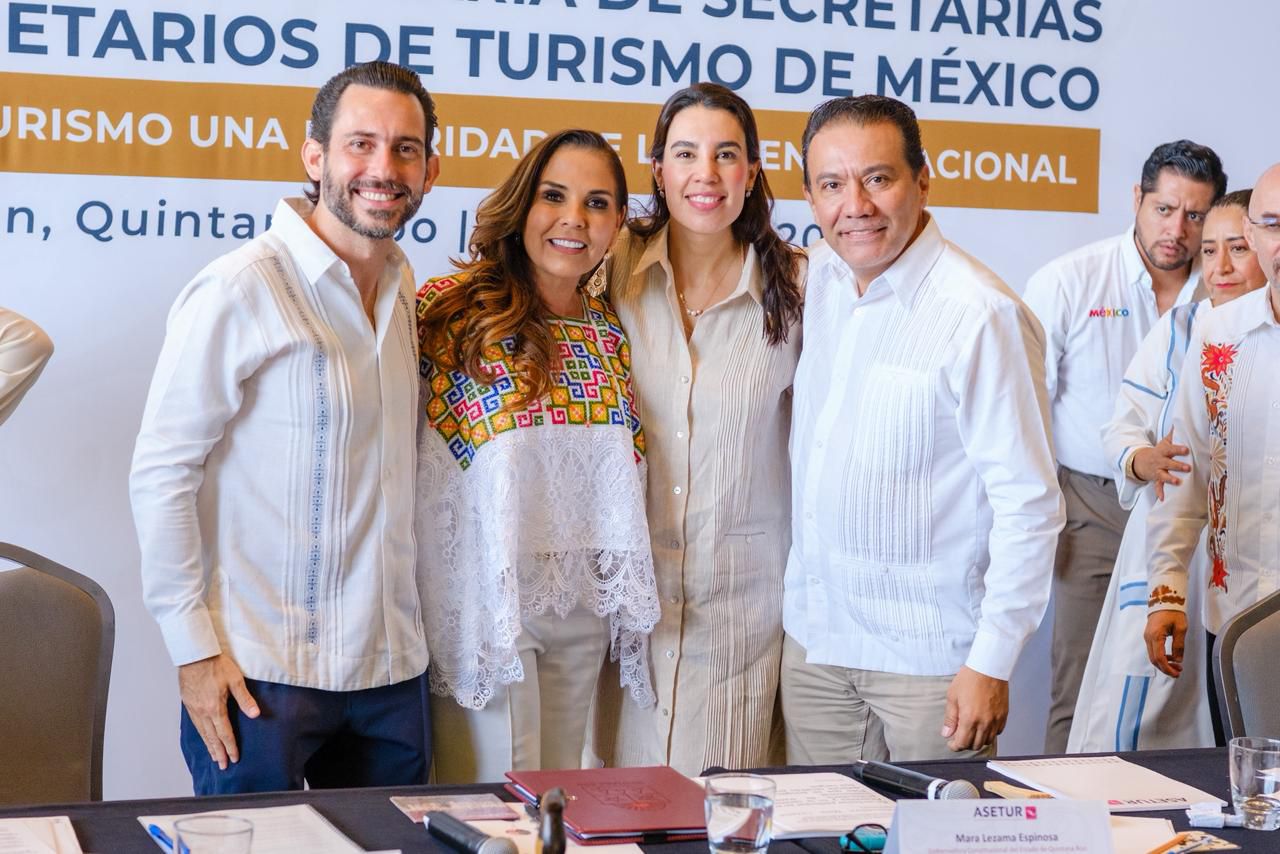 Fotografía En Plano Medio De Cuatro Funcionarios Sonriendo En Un Evento Oficial. De Izquierda A Derecha Aparecen Bernardo Cueto Riestra, La Gobernadora Mara Lezama Espinosa Vistiendo Una Blusa Blanca Con Bordados Tradicionales Coloridos Y Encaje, Josefina Rodríguez Zamora (Titular De Sectur) Y Roberto Monroy García. Todos Visten Ropa Formal Clara, Ideal Para Clima Cálido. Al Fondo Se Observa Una Lona Institucional Azul Y Blanca Con Letras Grandes Que Forman Parte Del Título Del Evento De La Asociación De Secretarios De Turismo De México.