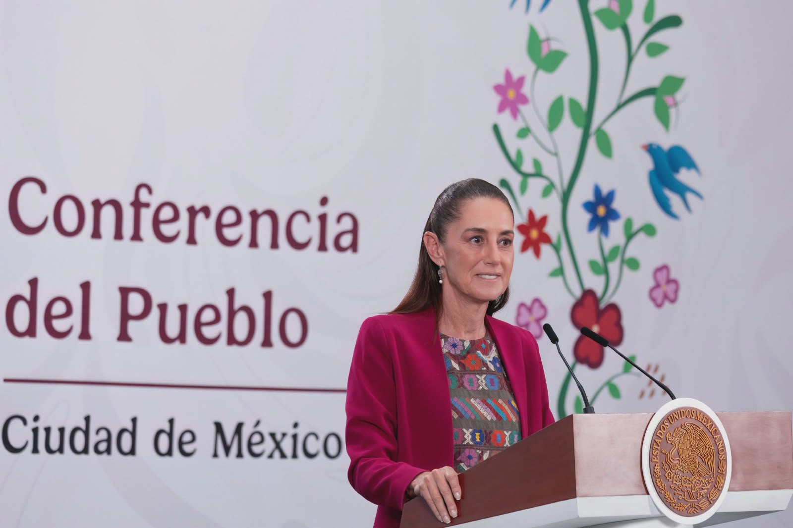 La Presidenta De México, Claudia Sheinbaum Pardo, Durante La Conferencia &Quot;Las Mañaneras Del Pueblo&Quot; En La Ciudad De México. Viste Un Saco Color Fucsia Y Una Blusa Con Bordados Tradicionales. Al Fondo Se Lee &Quot;Conferencia Del Pueblo&Quot; Junto A Una Ilustración Floral De Estilo Artesanal.