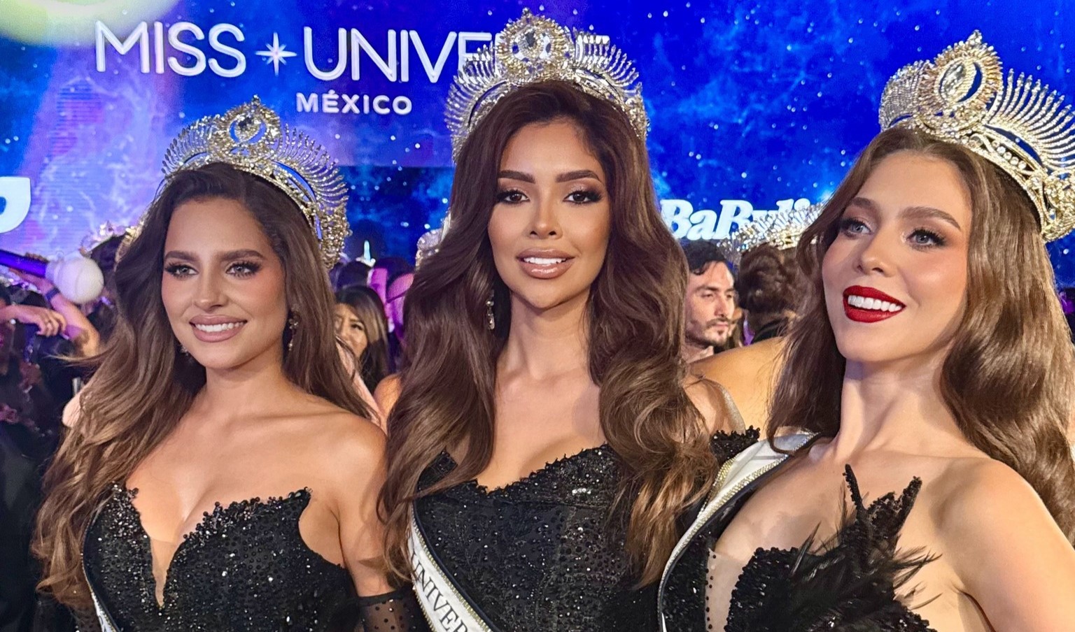 Fotografía En Plano Medio De Tres Reinas De Belleza Posando Juntas E Iluminadas En Un Escenario. Todas Lucen Cabello Largo Castaño Con Ondas, Maquillaje Glamoroso Y Grandes Coronas Plateadas E Intrincadas Sobre Sus Cabezas. La Mujer De La Izquierda Sonríe Con Un Vestido Negro Brillante De Escote Corazón; La Mujer Del Centro, Con Una Banda Blanca Que Dice &Quot;Universe&Quot;, Mira Al Frente Con Una Sonrisa Sutil Vistiendo Un Vestido Negro Con Pedrería; La Mujer De La Derecha Sonríe Ampliamente Mostrando Labios Rojos, Luciendo Un Atuendo Negro Con Detalles De Plumas. Al Fondo Se Aprecia Una Pantalla Azul Brillante Con Estrellas Que Muestra El Texto &Quot;Miss Universe México&Quot;, Además De Siluetas Desenfocadas De Público Y Fotógrafos.