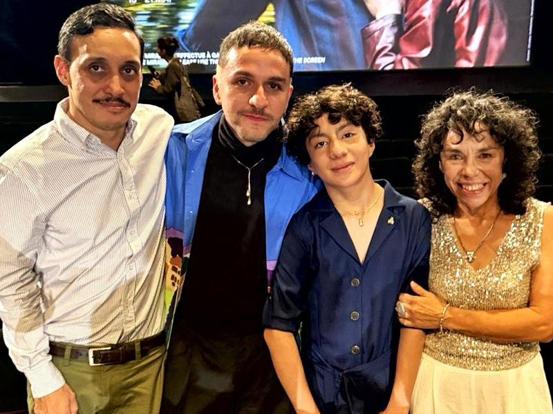 Fotografía Grupal En Plano Medio Que Muestra A Cuatro Integrantes Del Equipo Del Filme &Quot;Seis Meses En El Edificio Rosa Con Azul&Quot; Posando Juntos En El Interior De Una Sala De Cine En Cannes. De Izquierda A Derecha: Un Hombre Con Camisa De Rayas Claras Y Pantalón Verde Oliva; El Director Bruno Santamaría Razo Vistiendo Una Camisa Azul Abierta Sobre Una Playera Negra De Cuello Alto; El Joven Protagonista Jade Reyes Con Un Conjunto Azul Marino De Cuello En V; Y Una Mujer Sonriente Con Una Blusa Brillante De Lentejuelas Doradas Y Falda Blanca. El Grupo Posa Abrazado Y Sonriendo De Manera Cálida Hacia La Cámara, Con La Pantalla De Proyección Y Las Butacas Del Teatro Oscurecidas Al Fondo.