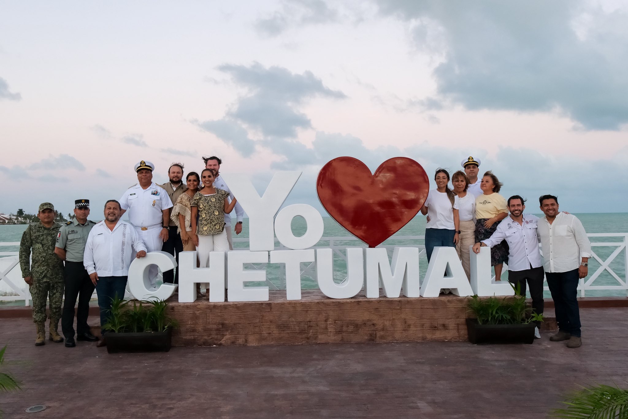 Fotografía Grupal De Autoridades Civiles Y Mandos Militares Posando Junto Al Parador Fotográfico De Letras Monumentales Blancas Que Forman La Frase &Quot;Yo Amo Chetumal&Quot;, Coronado Por Un Gran Corazón Rojo. Al Fondo Se Aprecia El Mar De La Bahía De Chetumal Bajo Un Cielo Nublado Durante El Atardecer, En El Entorno Del Recién Inaugurado Portal Marítimo De Chetumal.