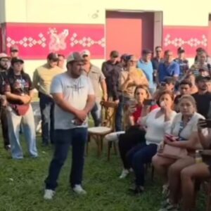 El Gobernador Javier May Rodríguez, Vistiendo Una Camisa Blanca De Manga Larga Y Pantalón De Mezclilla, Se Encuentra De Pie A La Izquierda, Dirigiéndose Con Semblante Serio A Un Grupo De Aproximadamente 40 Personas Sentadas Y De Pie En Una Zona De Césped.