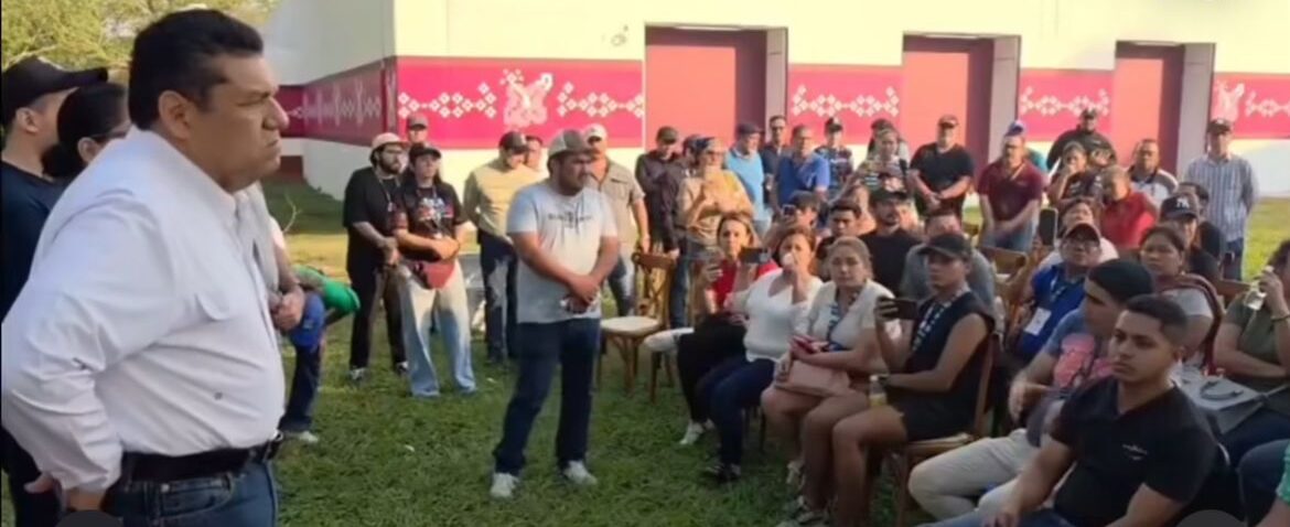 El Gobernador Javier May Rodríguez, Vistiendo Una Camisa Blanca De Manga Larga Y Pantalón De Mezclilla, Se Encuentra De Pie A La Izquierda, Dirigiéndose Con Semblante Serio A Un Grupo De Aproximadamente 40 Personas Sentadas Y De Pie En Una Zona De Césped.