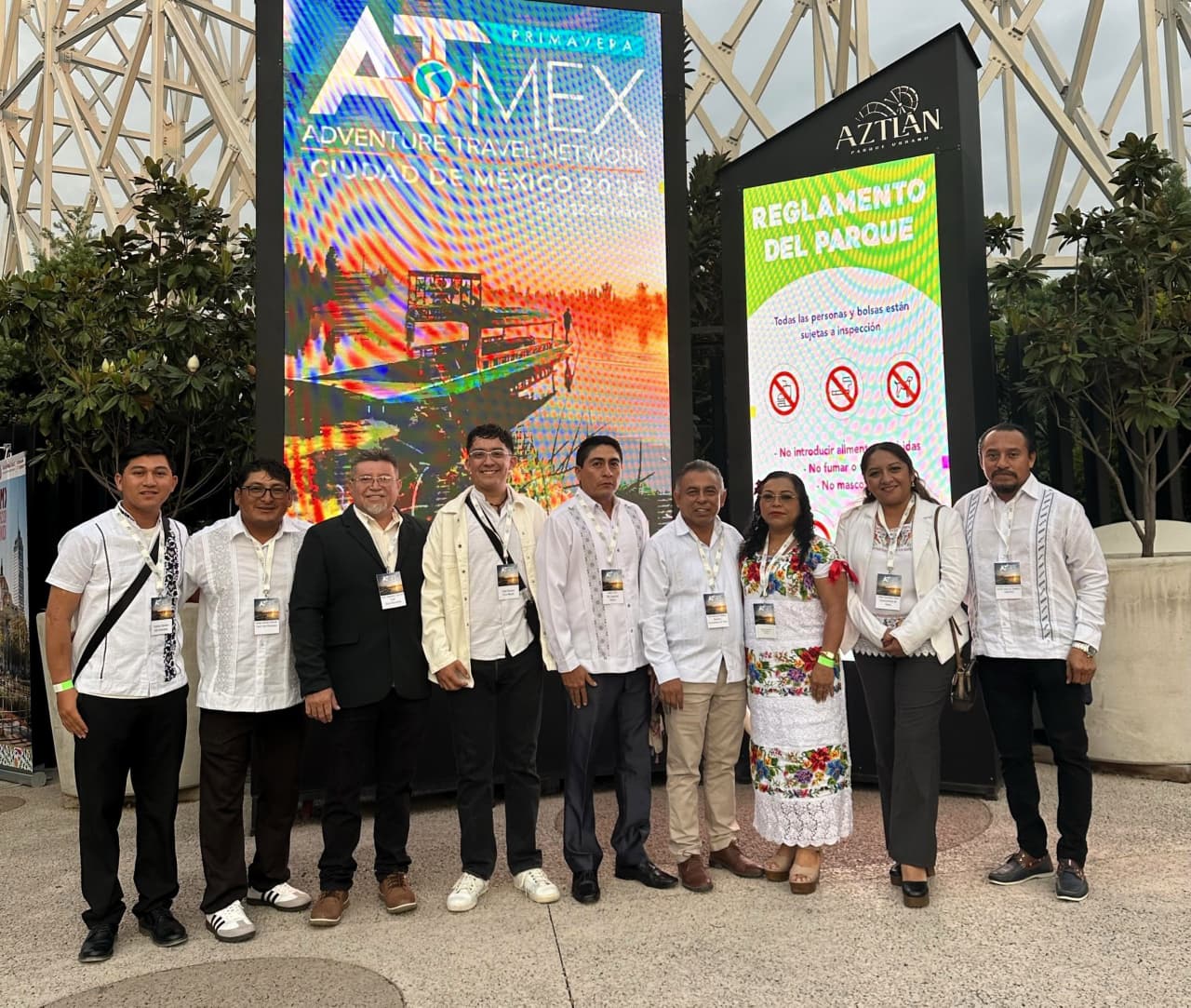 Turismo de aventura en yucatán destaca en atmex primavera 2026 la revista del sureste mx Un Grupo De Nueve Integrantes De La Delegación De Yucatán Posa Sonriente En Fila Para Una Fotografía Grupal Al Aire Libre. Visten Camisas Blancas, Guayaberas Tradicionales Y Trajes Regionales Bordados, Portando Gafetes Oficiales Del Evento. Al Fondo Se Aprecian Dos Grandes Pantallas Led, Una Con El Logotipo De Atmex Primavera Ciudad De México Y Otra Con El Reglamento Del Parque Urbano Aztlán, Flanqueadas Por Plantas Decorativas Y Una Estructura Metálica Blanca.
