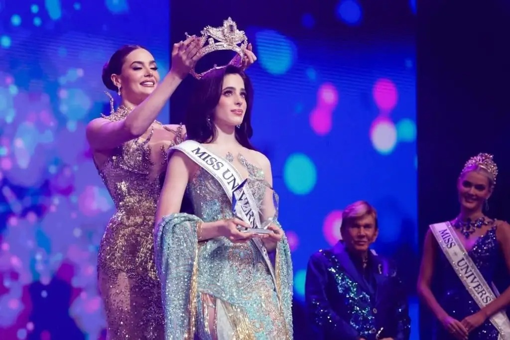Fotografía Del Momento En Que Fátima Bosch, Miss Universo 2025, Coloca La Corona A Su Sucesora Sobre Un Escenario Iluminado. La Nueva Reina Viste Un Elegante Vestido De Gala Azul Claro Con Pedrería Y Porta La Banda De Miss Universe. Al Fondo Se Observa A Osmel Sousa Y Otra Finalista En Un Ambiente De Celebración Con Luces De Colores.