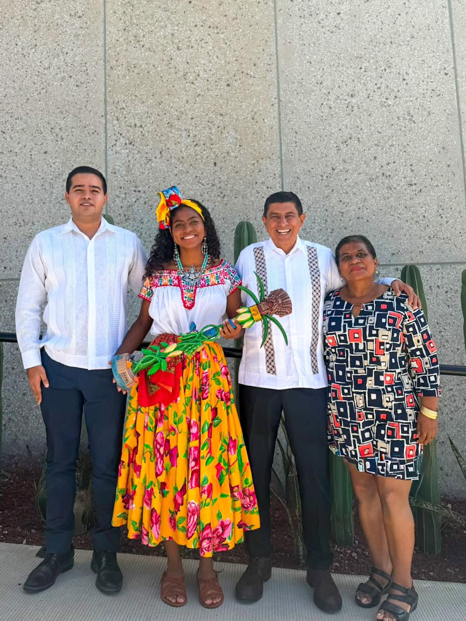 Conoce a enid azucena torres, la diosa centéotl 2026 que presidirá las fiestas de la guelaguetza la revista del sureste mx Fotografía Grupal De Cuerpo Completo En El Exterior. Al Centro, Enid Azucena Torres Agustiniano Sonríe Portando La Vestimenta Tradicional De Pinotepa Nacional, Que Incluye Una Falda Larga Amarilla Con Flores Rosadas, Blusa Blanca Bordada, Collares Tradicionales Y Un Tocado Colorido En El Cabello, Mientras Sostiene El Cetro Ceremonial De La Diosa Centéotl. A Su Izquierda, Un Joven Viste Guayabera Blanca Y Pantalón Oscuro.