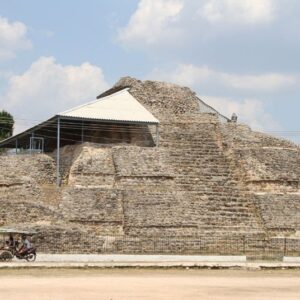 Acanceh: la impresionante pirámide maya que quedó en plena zona urbana de yucatán la revista del sureste mx Fotografía En Plano General Y Ángulo Contrapicado De La Pirámide De Los Mascarones En Acanceh, Yucatán. La Estructura De Piedra Caliza Muestra Sus Cuerpos Escalonados Y Desniveles Protegidos En La Esquina Superior Izquierda Por Una Estructura Con Techo De Lámina. Al Frente, Una Reja Metálica De Color Negro Corre A Lo Largo De Una Banqueta Blanca Que Delimita La Calle Pavimentada En Primer Plano. Varios Cables De Energía Eléctrica Cruzan Horizontalmente Toda La Toma Sobre Un Fondo De Cielo Azul Despejado Con Pocas Nubes.