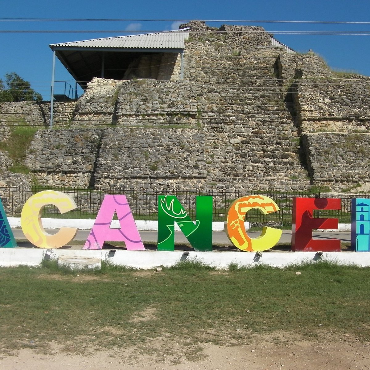 Acanceh: la impresionante pirámide maya que quedó en plena zona urbana de yucatán la revista del sureste mx Fotografía A Nivel De Piso Que Muestra Las Letras Turísticas Monumentales Y Coloridas Que Forman La Palabra &Quot;Acanceh&Quot;, Ubicadas Sobre Una Base Blanca Con Césped Al Frente. En La Letra &Quot;N&Quot; De Color Verde Destaca La Silueta De Un Venado. Justo Detrás De Las Letras Y Cruzando Una Calle Con Reja Metálica, Se Eleva La Pirámide De Los Mascarones, Una Estructura Arqueológica Maya De Piedra Con Niveles Escalonados Que Cuenta Con Una Techumbre De Lámina Gris En Su Sección Superior Izquierda Para Proteger Los Vestigios.