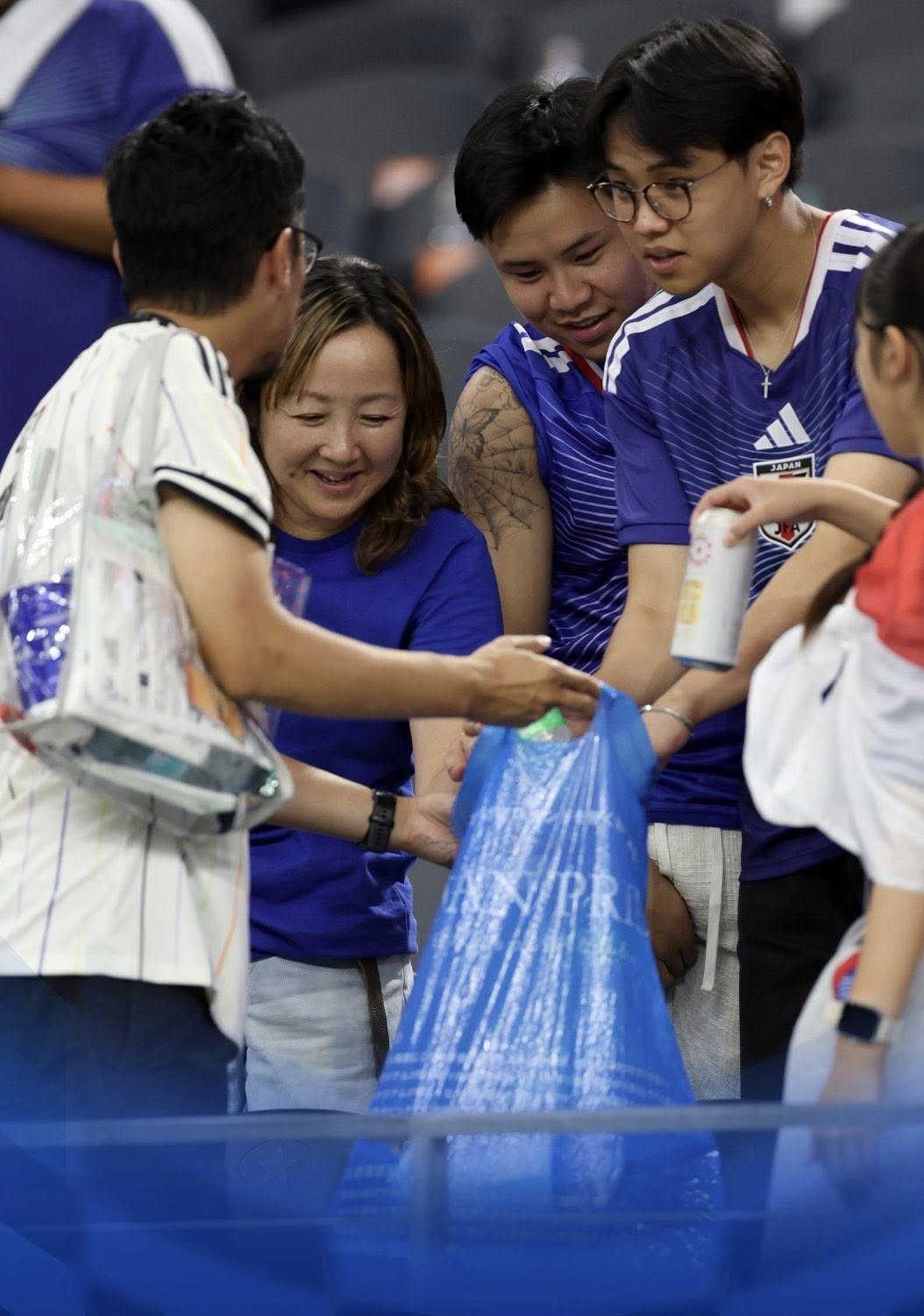 Lección de civismo en el mundial 2026: aficionados de japón limpian el estadio tras empate ante países bajos la revista del sureste mx Fotografía En Plano Medio Vertical De Un Grupo De Aficionados Japoneses Colaborando En La Limpieza De Las Gradas De Un Estadio. Al Centro, Varias Personas Sostienen Una Gran Bolsa De Plástico Azul Translúcido Mientras Introducen Botellas De Plástico Y Latas Vacías De Aluminio. Dos De Los Jóvenes Visten La Playera Oficial De Fútbol De La Selección De Japón En Color Azul Con Detalles Blancos.