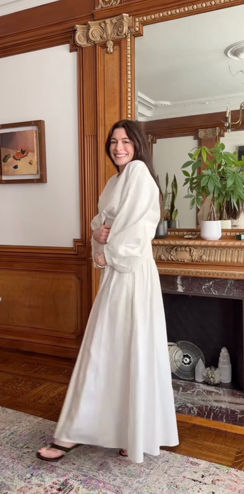 ¡sorpresa en hollywood! Anne hathaway anuncia que está embarazada a los 43 años y espera a su tercer hijo la revista del sureste mx Una Fotografía De Cuerpo Completo De La Actriz Anne Hathaway Sonriendo Alegremente En El Interior De Una Habitación Con Diseño Clásico. Ella Está De Perfil, Vistiendo Un Vestido Largo Y Holgado De Color Blanco Con Mangas Abullonadas, Y Lleva Puestas Sandalias Oscuras. Sus Manos Descansan Suavemente Sobre Su Incipiente Vientre De Embarazo. El Fondo Destaca Por Detalles Elegantes De Madera Tallada En La Pared, Un Gran Espejo Con Marco Ornamental, Una Chimenea De Mármol Oscuro Y Plantas Decorativas En Macetas Blancas Sobre La Repisa.