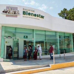Una Fotografía Que Muestra Una Sucursal Moderna Del Banco Del Bienestar. Varias Personas, En Su Mayoría Adultos Mayores, Hacen Fila En El Exterior, Junto A La Rampa De Acceso. Una Mujer Está Usando Un Cajero Automático En La Entrada. El Edificio Es Blanco Con Fachada De Cristal Y Tiene El Letrero Del Banco Con La Frase &Amp;Quot;El Banco De Los Mexicanos&Amp;Quot;.
