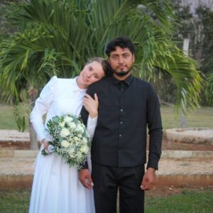 Retrato De Pie En Plano Medio De Los Recién Casados Katarina Y Camilo En Hopelchén, Campeche. A La Izquierda, La Joven De La Comunidad Menonita Sonríe Sutilmente Mientras Apoya Su Cabeza En El Hombro De Su Esposo Y Sostiene Un Gran Ramo De Rosas Blancas Y Follaje Verde; Viste Su Vestido De Novia Tradicional Blanco Con Mangas Largas. A La Derecha, El Joven Campechano Posa Con Semblante Serio, Vistiendo Una Camisa Negra Y Un Chaleco Abotonado Del Mismo Color. El Fondo Muestra Un Entorno Natural Al Aire Libre Con Pasto, Jardineras De Piedra Baja Y Una Palmera Pequeña Directamente Detrás De La Pareja.