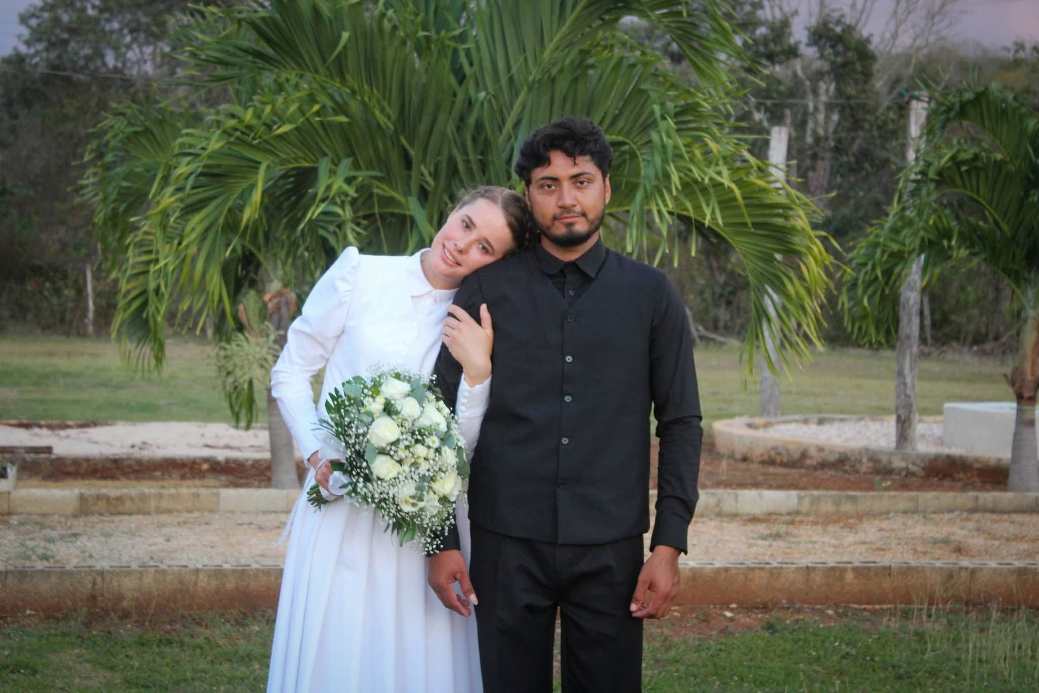 Boda menonita y chenera en hopelchén une dos culturas en campeche la revista del sureste mx Retrato De Pie En Plano Medio De Los Recién Casados Katarina Y Camilo En Hopelchén, Campeche. A La Izquierda, La Joven De La Comunidad Menonita Sonríe Sutilmente Mientras Apoya Su Cabeza En El Hombro De Su Esposo Y Sostiene Un Gran Ramo De Rosas Blancas Y Follaje Verde; Viste Su Vestido De Novia Tradicional Blanco Con Mangas Largas. A La Derecha, El Joven Campechano Posa Con Semblante Serio, Vistiendo Una Camisa Negra Y Un Chaleco Abotonado Del Mismo Color. El Fondo Muestra Un Entorno Natural Al Aire Libre Con Pasto, Jardineras De Piedra Baja Y Una Palmera Pequeña Directamente Detrás De La Pareja.