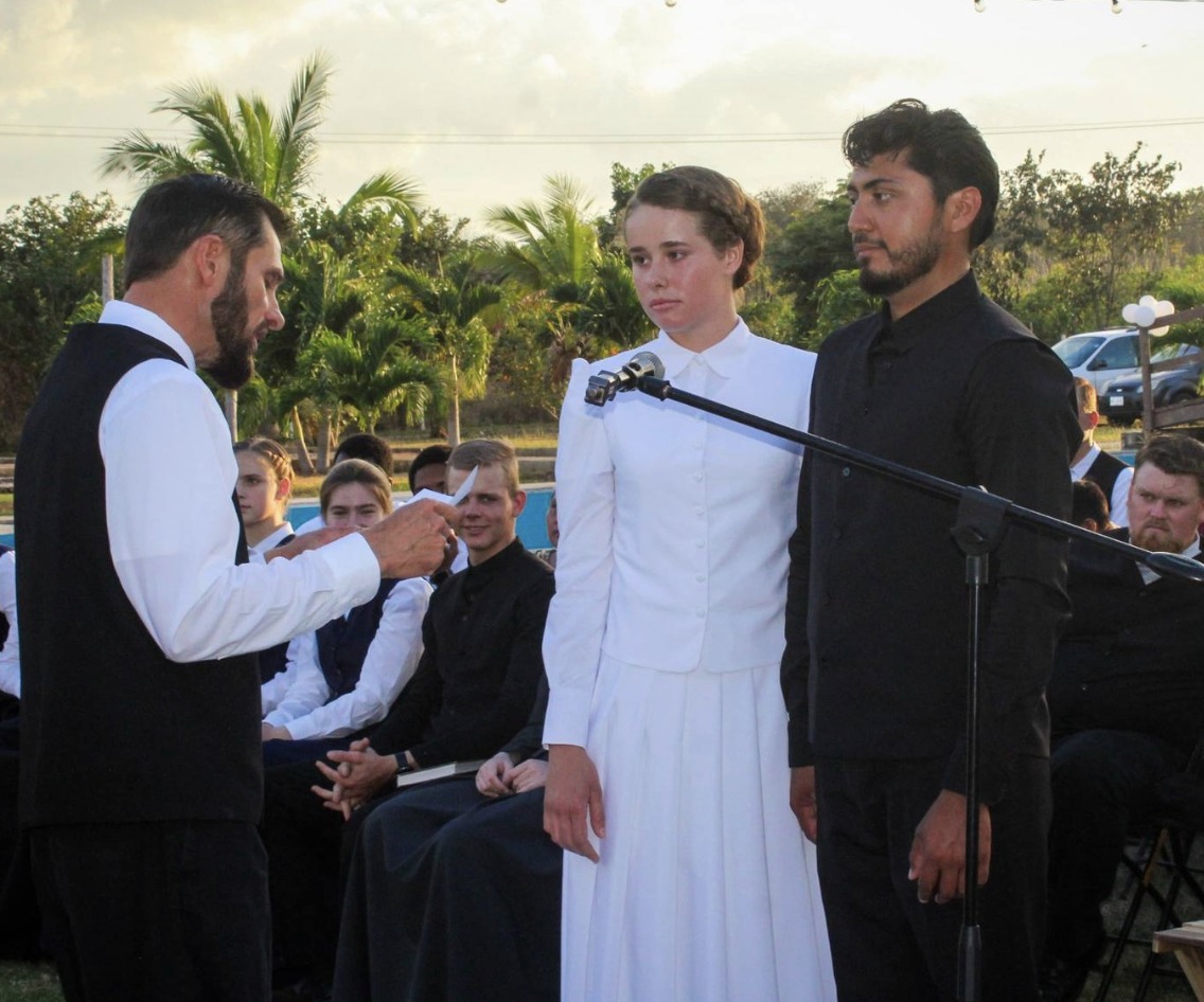 Fotografía En Plano Medio De Una Ceremonia Matrimonial Al Aire Libre En Hopelchén, Campeche. En El Centro, Una Joven De La Comunidad Menonita Viste Un Traje De Novia Tradicional Blanco, De Manga Larga Y Cuello Cerrado, Con El Cabello Recogido; Junto A Ella, Un Joven Originario De La Región Viste Una Camisa Y Chaleco Negros. Frente A Ellos, Un Ministro Con Chaleco Negro Y Camisa Blanca Lee Un Documento. Al Fondo, Se Observa A Los Invitados Sentados, Vegetación Con Palmeras Bajo Una Luz De Atardecer, Un Micrófono De Pedestal Frente A La Pareja Y Un Vehículo Estacionado A La Distancia.