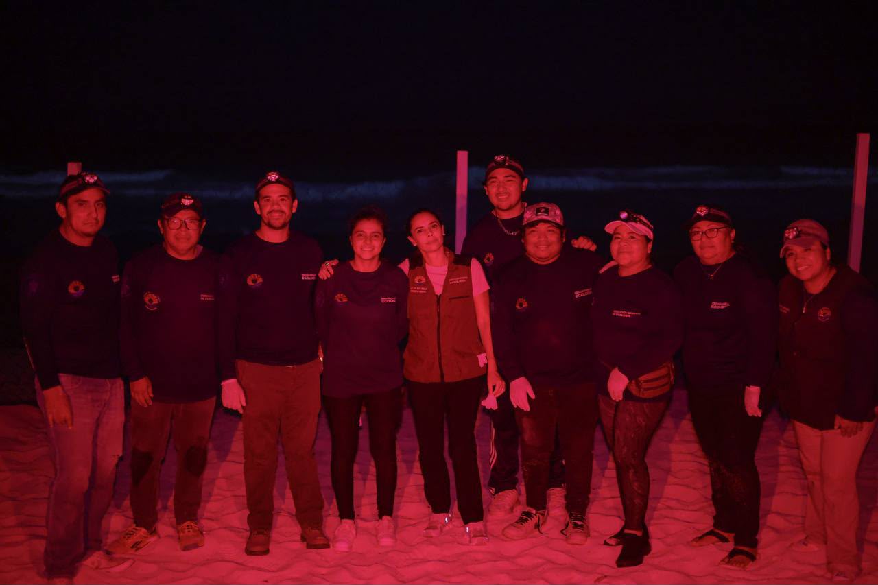 Fotografía Nocturna En Una Playa De Cancún Protegida, Donde El Personal Del Programa De Conservación Posa En Grupo Bajo Luz Roja. El Equipo, Vestido Con Uniformes Azules Con Logotipos Y Chalecos, Está Sobre La Arena Con La Orilla Oscura Y Postes De Cerramiento De Nidos.