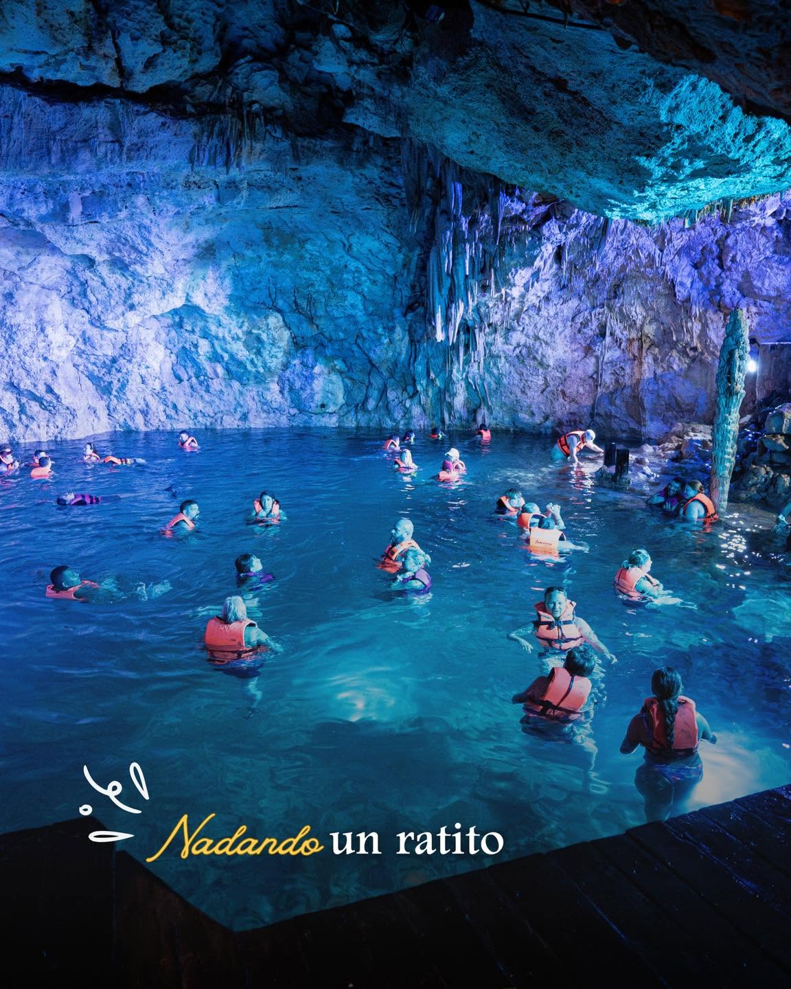 Descripción: Fotografía De Ángulo Medio Tomada Desde Una Plataforma De Madera A Nivel Del Agua Que Captura Una Escena De Nado Grupal En La Caverna Subterránea De Los Cenotes De Santa Rosa En Homún. Un Grupo Numeroso De Más De Quince Personas, Todas Vistiendo Chalecos Salvavidas De Color Naranja Brillante Para Seguridad, Está Disperso Nadando En Las Aguas Cristalinas Y Profundas Que Reflejan Intensos Tonos Turquesa Y Azul Debido A La Iluminación Artificial Instalada En La Cueva.