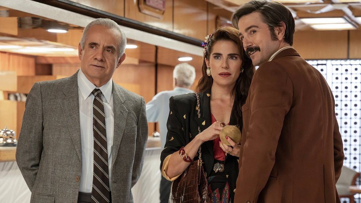 Se Observa Un Fotograma De La Película Ambientado En La Década De Los Ochenta Dentro De Lo Que Parece Ser El Vestíbulo O Recepción De Un Hotel. A La Derecha, El Actor Diego Luna, Con Cabello Peinado Hacia Atrás, Bigote Espeso Y Un Saco Color Marrón, Mira De Reojo Hacia La Izquierda Con Expresión Atenta. Junto A Él, Una Mujer Con Cabello Largo Castaño Adornado Con Pedrería, Aretes Grandes Y Un Saco Negro Con Detalles Dorados, Sostiene Una Esfera Dorada Con Ambas Manos Mientras Observa Fijamente Hacia El Mismo Punto. A La Izquierda, El Actor Daniel Giménez Cacho