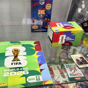 Fotografía Tomada Desde Un Ángulo Ligeramente Elevado Que Muestra El Interior De Una Vitrina De Cristal Transparente Con Diversos Coleccionables De La Marca Panini. En La Parte Inferior Izquierda Destaca El Álbum De Pasta Blanda Oficial De La &Amp;Quot;Fifa World Cup 2026&Amp;Quot;, Con Un Llamativo Diseño Gráfico En Tonos Verdes, Amarillos Y Rojos, Mostrando El Trofeo Dorado De La Copa Del Mundo En El Centro Y Una Etiqueta Que Dice &Amp;Quot;Somos México&Amp;Quot;.
