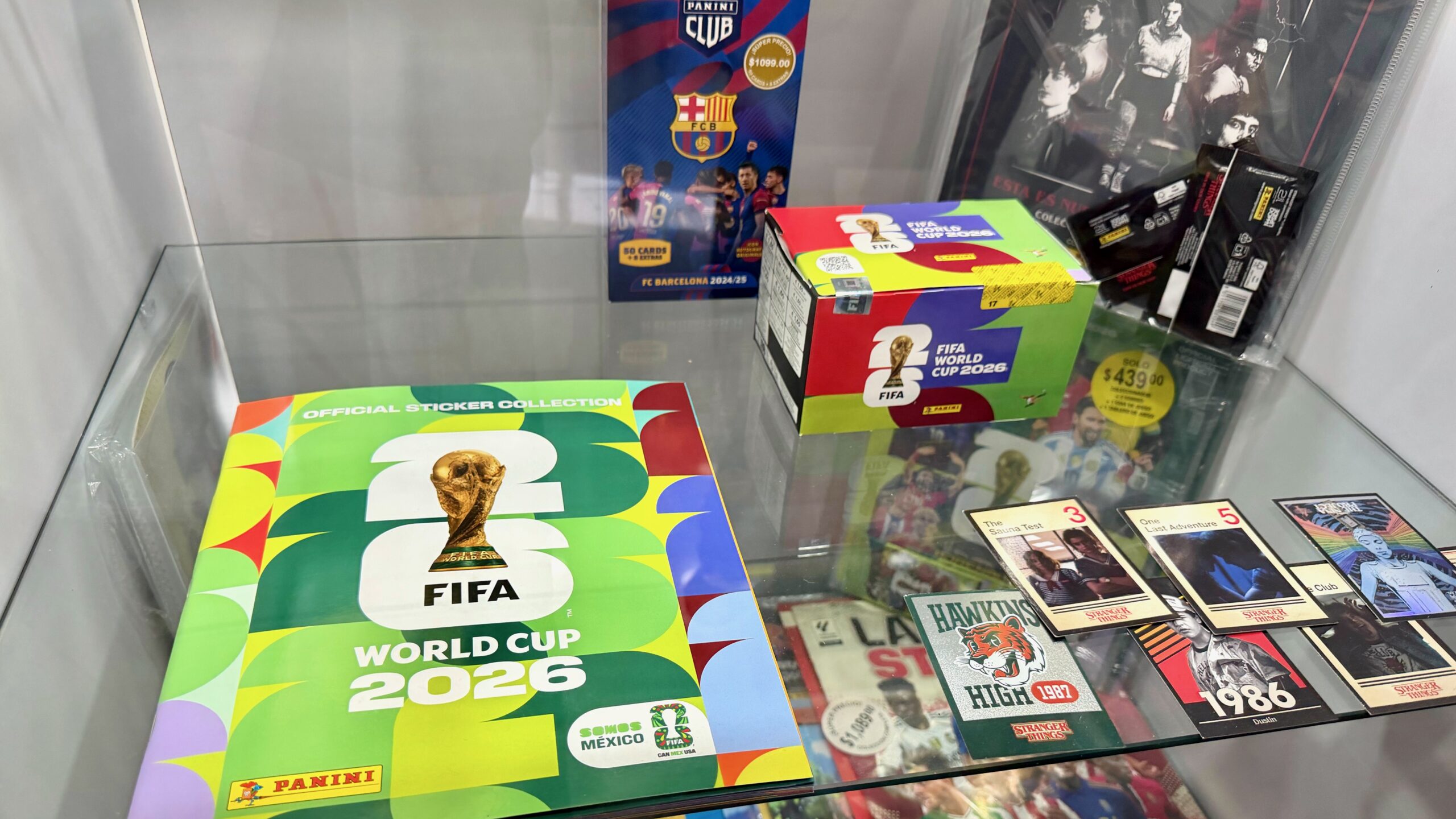 Fotografía Tomada Desde Un Ángulo Ligeramente Elevado Que Muestra El Interior De Una Vitrina De Cristal Transparente Con Diversos Coleccionables De La Marca Panini. En La Parte Inferior Izquierda Destaca El Álbum De Pasta Blanda Oficial De La &Quot;Fifa World Cup 2026&Quot;, Con Un Llamativo Diseño Gráfico En Tonos Verdes, Amarillos Y Rojos, Mostrando El Trofeo Dorado De La Copa Del Mundo En El Centro Y Una Etiqueta Que Dice &Quot;Somos México&Quot;.