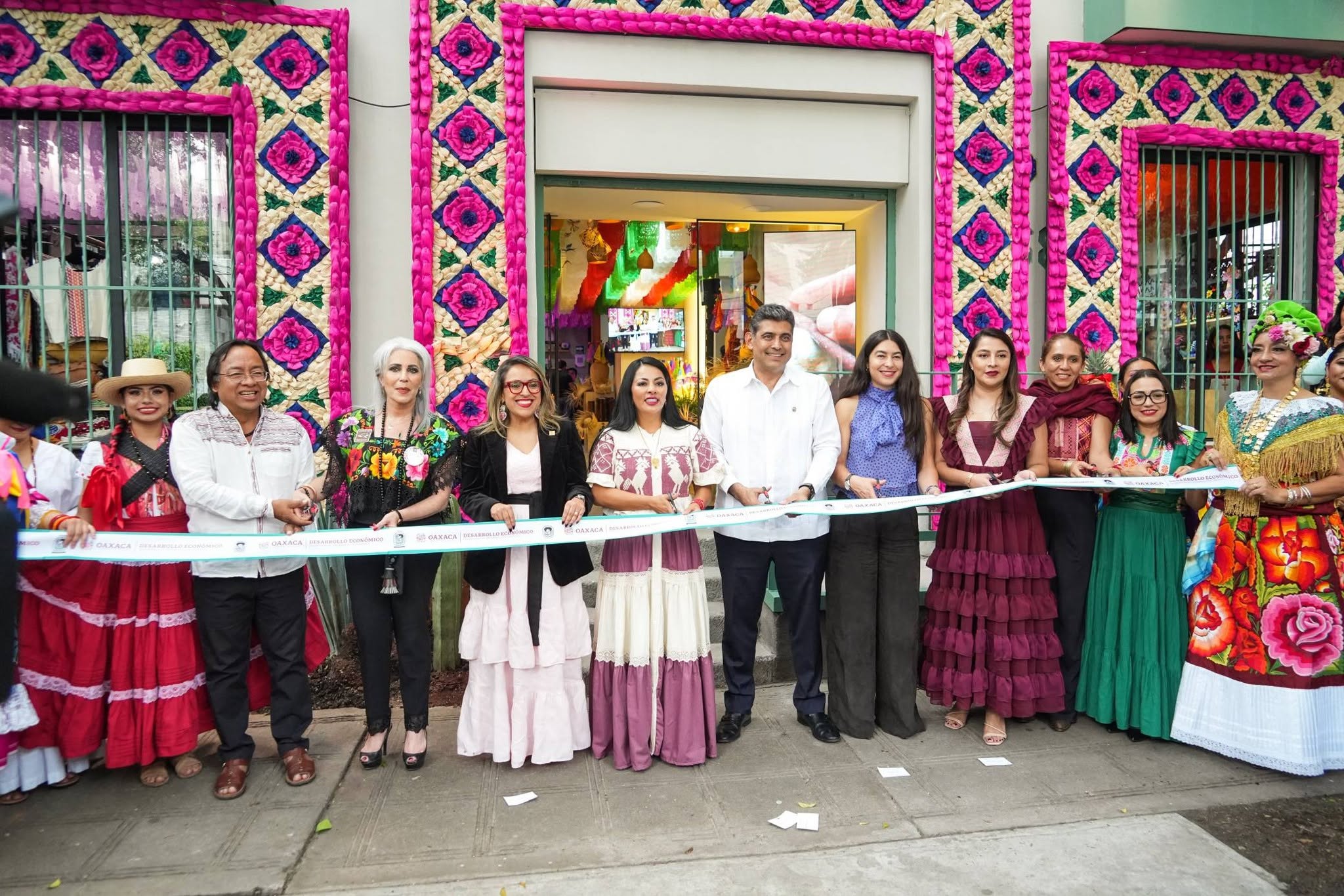 Con toda su cultura, gastronomía e historia, inauguran la "casa hecho en oaxaca" en la ciudad de méxico para promover la riqueza de sus ocho regiones la revista del sureste mx Una Fotografía De Ángulo Medio A Nivel De Los Ojos Captura A Un Grupo De Personas De Pie En Una Línea En Una Acera De Hormigón Frente A La Entrada De Una Tienda. Los Miembros Del Grupo, Una Mezcla De Hombres Y Mujeres, Sonríen Mientras Cortan Una Larga Cinta Azul Claro. Varios Miembros Del Grupo Llevan Ropa Tradicional Mexicana Oaxaqueña, Como Blusas Bordadas Con Flores Y Faldas Completas De Color Rojo, Burdeos Y Verde, Mientras Que Otros Llevan Trajes Empresariales Modernos.