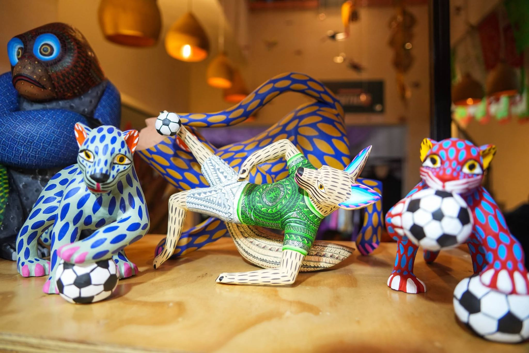 Con toda su cultura, gastronomía e historia, inauguran la "casa hecho en oaxaca" en la ciudad de méxico para promover la riqueza de sus ocho regiones la revista del sureste mx Una Toma De Ángulo Medio En Primer Plano Muestra Varias Tallas De Madera De Alebrijes Oaxaqueños Pintadas De Manera Colorida, Sentadas Sobre Una Superficie De Madera. La Figura Central Es Un Coyote O Perro Con Una Camiseta De Fútbol Verde Con Patrones Ornamentados Y Pantalones Cortos Blancos, En Una Pose Dinámica Mientras Patea Un Pequeño Balón De Fútbol.