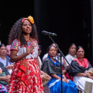 Conoce a enid azucena torres, la diosa centéotl 2026 que presidirá las fiestas de la guelaguetza la revista del sureste mx Enid Azucena Torres Agustiniano, Representante De Pinotepa Nacional, De Pie Frente A Un Micrófono En El Escenario Del Teatro Macedonio Alcalá Durante El Certamen Diosa Centéotl 2026. La Joven Oaxaqueña Viste La Indumentaria Tradicional De La Costa Con Falda Roja Floreada Y Blusa Blanca Con Bordados Coloridos, Luciendo Su Cabello Rizado Suelto Con Un Tocado Amarillo. Su Mano Derecha Descansa Sobre Su Pecho Mientras Emite Su Discurso. Al Fondo, Sentadas En El Escenario, Se Observan Varias De Las Demás Participantes Del Certamen Vistiendo Trajes Típicos De Diversas Regiones De Oaxaca. La Iluminación Destaca Su Participación Central