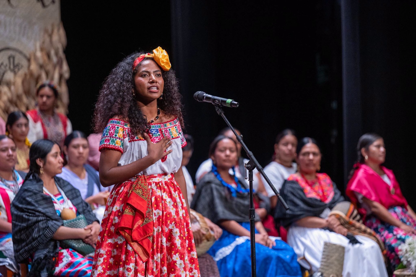 Conoce a enid azucena torres, la diosa centéotl 2026 que presidirá las fiestas de la guelaguetza la revista del sureste mx Enid Azucena Torres Agustiniano, Representante De Pinotepa Nacional, De Pie Frente A Un Micrófono En El Escenario Del Teatro Macedonio Alcalá Durante El Certamen Diosa Centéotl 2026. La Joven Oaxaqueña Viste La Indumentaria Tradicional De La Costa Con Falda Roja Floreada Y Blusa Blanca Con Bordados Coloridos, Luciendo Su Cabello Rizado Suelto Con Un Tocado Amarillo. Su Mano Derecha Descansa Sobre Su Pecho Mientras Emite Su Discurso. Al Fondo, Sentadas En El Escenario, Se Observan Varias De Las Demás Participantes Del Certamen Vistiendo Trajes Típicos De Diversas Regiones De Oaxaca. La Iluminación Destaca Su Participación Central