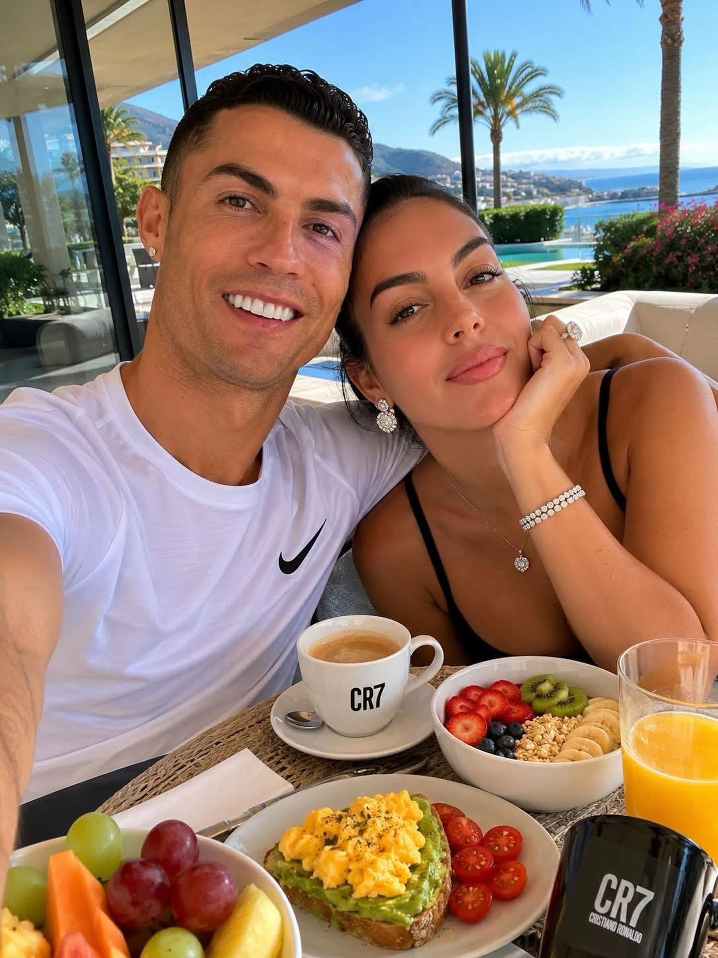 Una Selfie En Plano Medio De Cristiano Ronaldo Y Georgina Rodríguez Sentados Juntos Durante Un Desayuno Al Aire Libre. A La Izquierda De La Imagen, Cristiano Sonríe Frontalmente Vistiendo Una Playera Deportiva Blanca De Cuello Redondo Con Un Logotipo Negro.