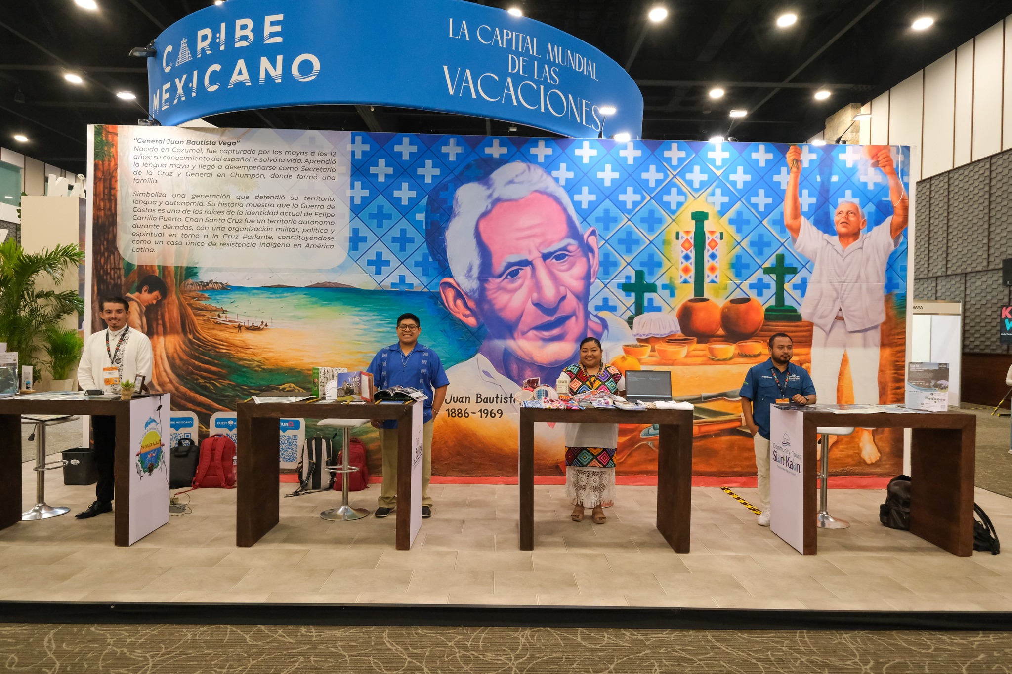 Conoce las 8 experiencias de turismo comunitario con las que maya ka’an y quintana roo deslumbran en k’íiwik 2026 la revista del sureste mx Se Muestra El Stand Promocional Del Caribe Mexicano Dentro De Una Feria Turística. Al Fondo, Un Gran Mural Colorido Ilustra El Rostro De Un Líder Indígena Con La Inscripción &Quot;Juan Bautista Vega 1886-1969&Quot;, Rodeado De Símbolos Tradicionales, Cruces Verdes, Un Paisaje Playero Y Un Texto Informativo Sobre La Resistencia Indígena En Felipe Carrillo Puerto.