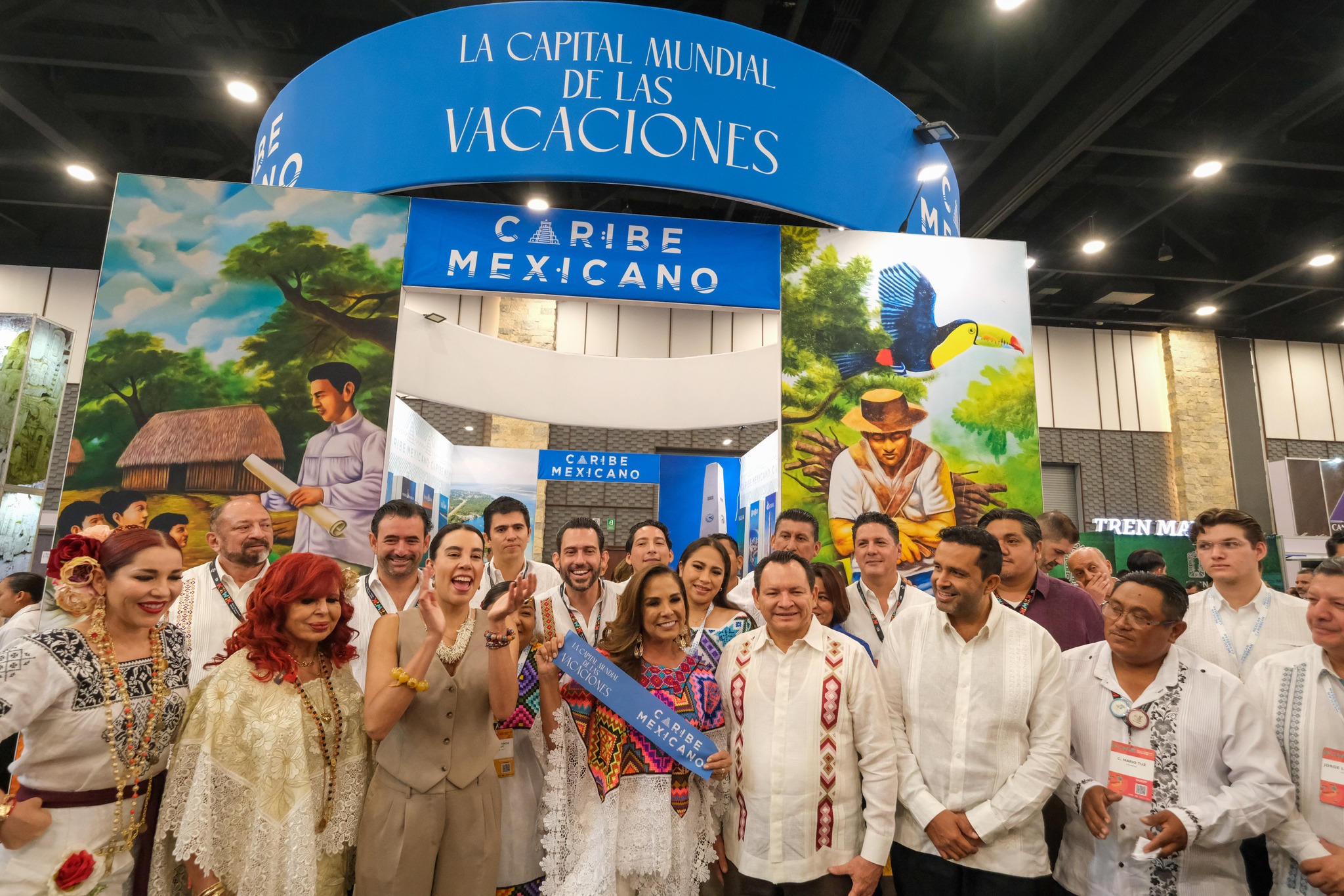 Un Grupo Numeroso De Funcionarios Públicos, Delegados Y Personas Con Vestimentas Tradicionales Posan Sonrientes Para Una Fotografía Grupal Frente Al Pabellón Del Caribe Mexicano. Al Centro, La Gobernadora Mara Lezama Viste Un Huipil Blanco Bordado Con Coloridos Patrones Geométricos Y Sostiene Un Listón Azul Con Letras Blancas Que Dice &Quot;Caribe Mexicano&Quot;. A Su Lado Izquierdo Se Encuentra El Gobernador Joaquín Díaz Mena